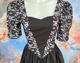 Puede incluir: Un vestido negro con escote cuadrado y mangas cortas abullonadas. Las mangas y el corpiño presentan detalles de encaje blanco. El vestido tiene una falda amplia y se exhibe en un maniquí.