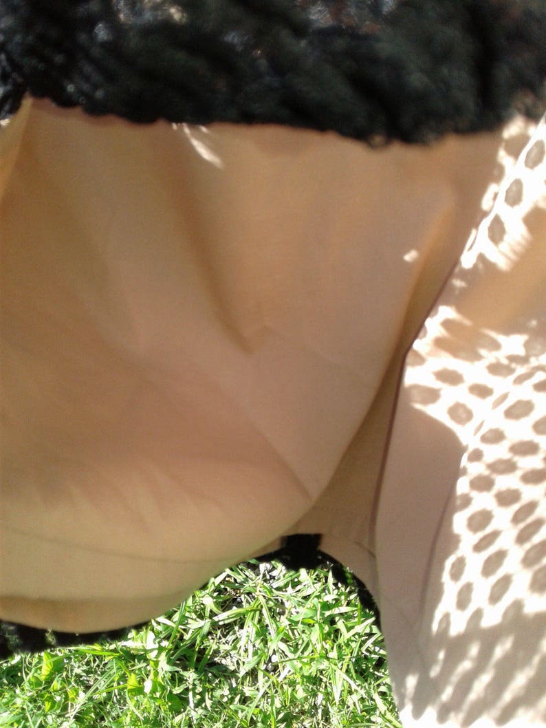 Puede incluir: Una falda beige con una superposici&oacute;n de encaje negro. La falda est&aacute; parcialmente abierta, revelando la tela beige debajo. La falda est&aacute; sobre un lecho de hierba verde.