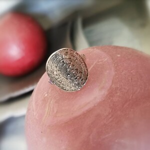 May include: A silver ring with a circular design featuring intricate details and a raised center. The ring is on a pink surface.