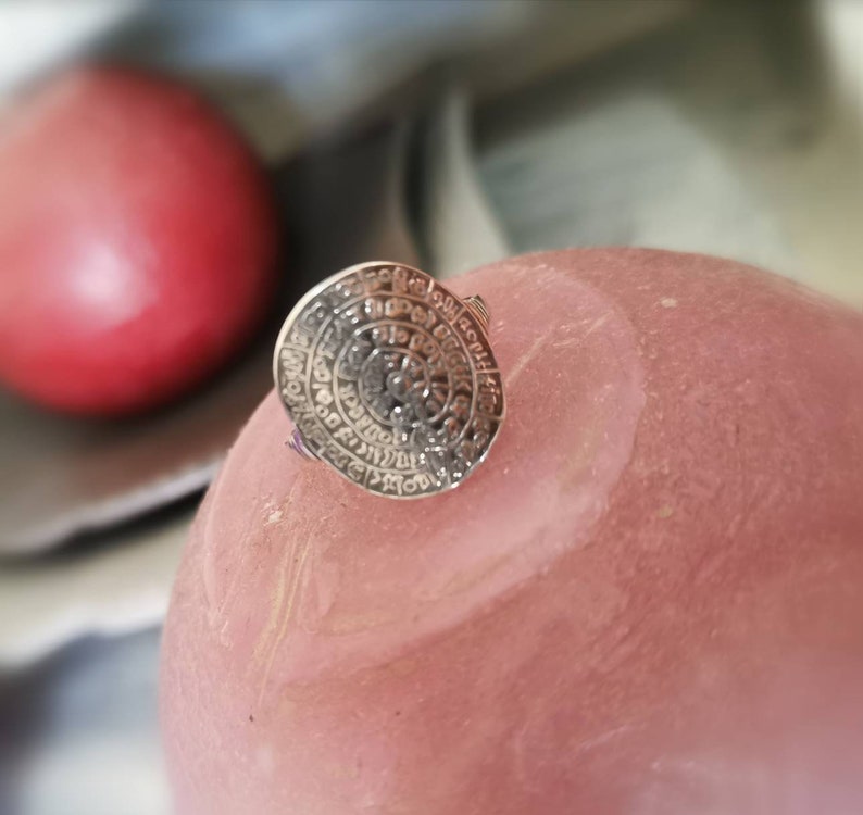 May include: A silver ring with a circular design featuring intricate details and a raised center. The ring is on a pink surface.