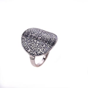 May include: A silver ring with a large, circular face featuring a spiral design and intricate markings. The ring is made of metal and has a polished finish.