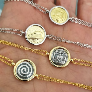 Könnte beinhalten: Vier goldene und silberne Armbänder mit verschiedenen Münzenförmigen Anhängern. Die Anhänger zeigen ein Spiralmuster, einen griechischen Tempel, eine geflügelte Kreatur und ein stilisiertes Design.