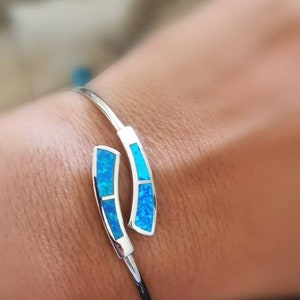 May include: A silver bangle bracelet with two blue opal accents. The bracelet is open and has a curved design.