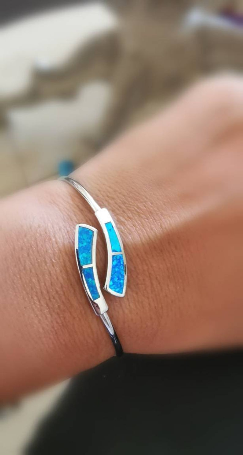 May include: A silver bangle bracelet with two blue opal accents. The bracelet is open and has a curved design.
