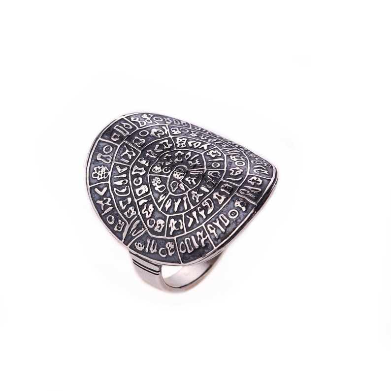 May include: A silver ring with a large, circular face featuring an intricate design of symbols and runes. The ring is made of a textured metal and has a slightly curved shape.