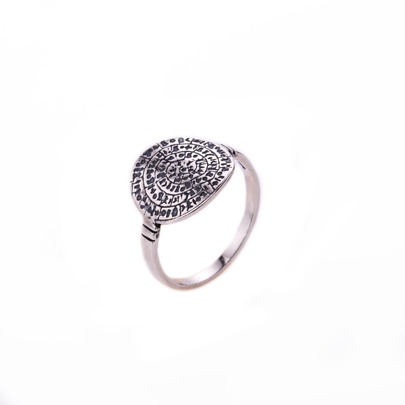 May include: A silver ring with a circular design featuring a pattern of small, intricate symbols. The ring is on a white background.