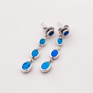 May include: Silver drop earrings with blue opal stones. Each earring has three oval, blue stones linked by a silver chain. The earrings have a post and backing. The opal stones have a bright blue colour. The earrings are a classic design.