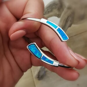 May include: A silver bangle bracelet with two blue opal accents. The bracelet is open and has a simple design.