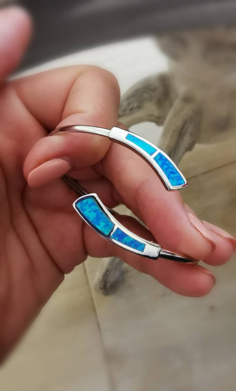 May include: A silver bangle bracelet with two blue opal accents. The bracelet is open and has a simple design.