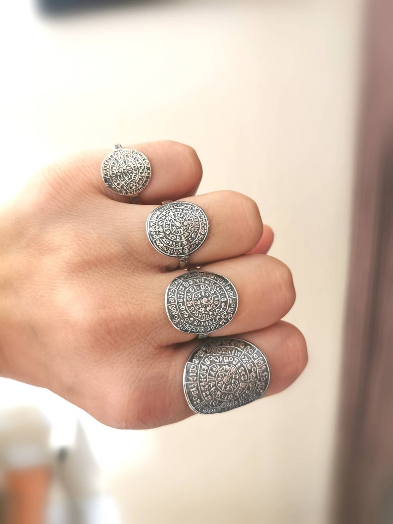 May include: Four silver rings with a circular design featuring intricate patterns and symbols. The rings are worn on a hand.