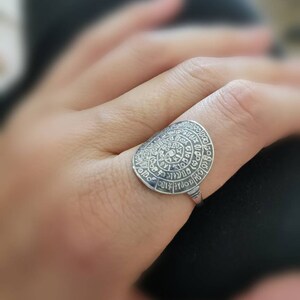 May include: A silver ring with a circular design featuring a spiral of text. The ring is worn on a finger.