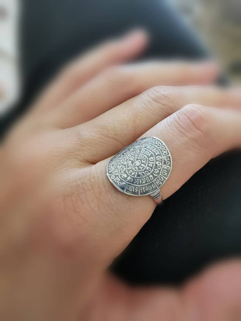 May include: A silver ring with a circular design featuring a spiral of text. The ring is worn on a finger.