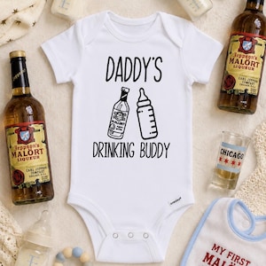 May include: A white short-sleeved baby bodysuit featuring the black text DADDY'S DRINKING BUDDY with a graphic illustration of a liquor bottle and a baby bottle. The design is centred on the chest of the garment, which has a round neckline.