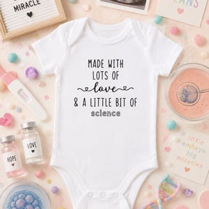 Może przedstawiać: Białe body niemowlęce z krótkim rękawem z czarnym nadrukiem "MADE WITH LOTS OF love & A LITTLE BIT OF science" na środku klatki piersiowej, z trzema zatrzaskami w dolnej części i klasycznym okrągłym dekoltem.