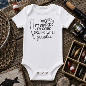 Peut inclure: Un body pour bébé blanc à manches courtes avec le texte noir "PACK MY DIAPERS I'M GOING FISHING WITH grandpa" accompagné d'une illustration graphique d'une canne à pêche et d'un petit poisson. Le vêtement a un col rond et trois boutons-pression.