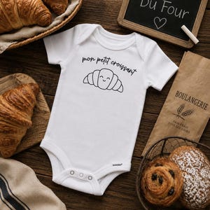 May include: A white short-sleeved baby bodysuit featuring a black line drawing of a smiling croissant and the text "mon petit croissant" printed on the chest. The garment has a round neckline and three metal snap closures at the bottom.