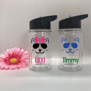 May include: Two clear plastic water bottles with black lids and flip-top spouts. Each bottle has a cartoon cat face with sunglasses and a name on the bottom. The left bottle has a pink bow and the name "Lexi". The right bottle has blue sunglasses and the name "Timmy".