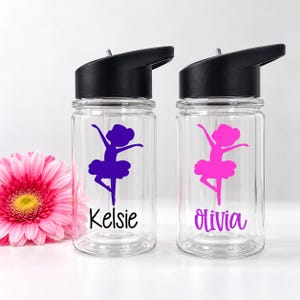 May include: Two clear plastic water bottles with black lids and straws. Each bottle features a ballerina silhouette graphic and a name printed in black script. One bottle has a purple graphic and the name Kelsie, the other a pink graphic and Olivia.