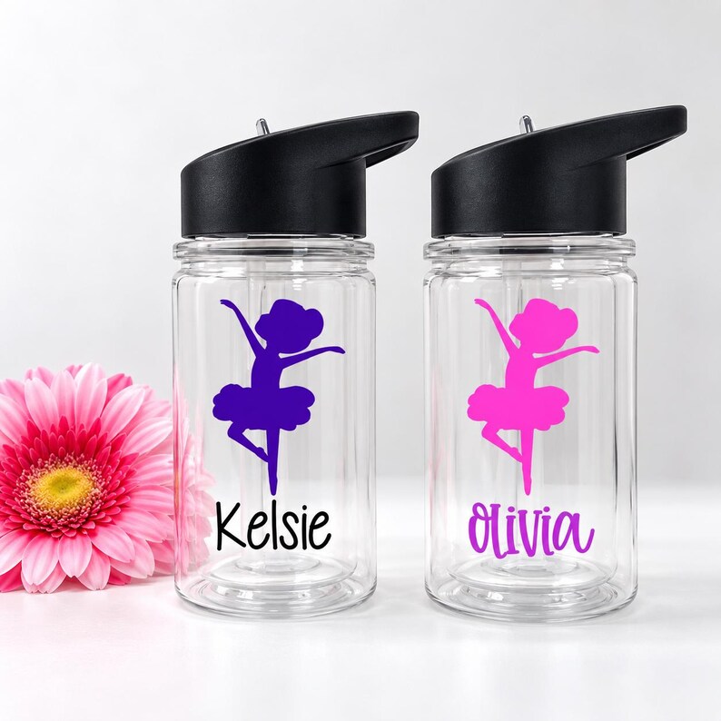 May include: Two clear plastic water bottles with black lids and straws. Each bottle features a ballerina silhouette graphic and a name printed in black script. One bottle has a purple graphic and the name Kelsie, the other a pink graphic and Olivia.