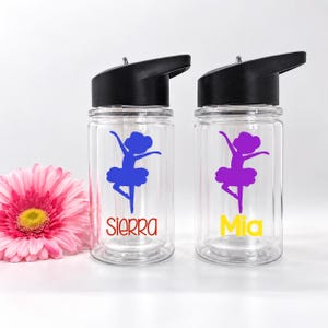 May include: Two clear plastic water bottles with black lids. Each bottle features a purple graphic of a ballerina in a tutu. One bottle has the name Sierra in red text, and the other has the name Mia in yellow text below the ballerina graphic.