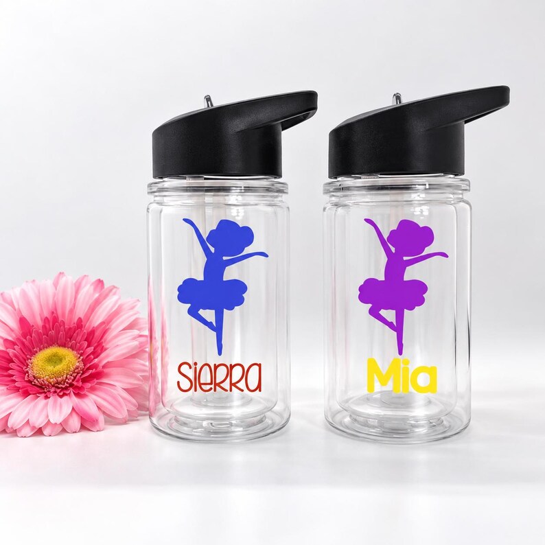 May include: Two clear plastic water bottles with black lids. Each bottle features a purple graphic of a ballerina in a tutu. One bottle has the name Sierra in red text, and the other has the name Mia in yellow text below the ballerina graphic.