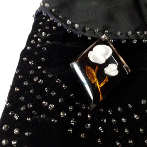 Könnte beinhalten: Eine klare, rautenförmige Brosche mit einem Design aus weißen gepressten Blumen. Die Brosche ist vor einem schwarzen Stoffhintergrund mit kleinen, klaren Strasssteinen platziert.