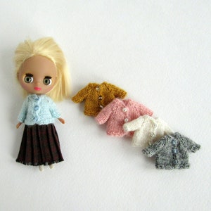 May include: A doll wearing a light blue knitted cardigan, a brown plaid skirt, and blonde hair. The doll is surrounded by five miniature knitted cardigans in various colors, including pink, white, gray, and mustard yellow.