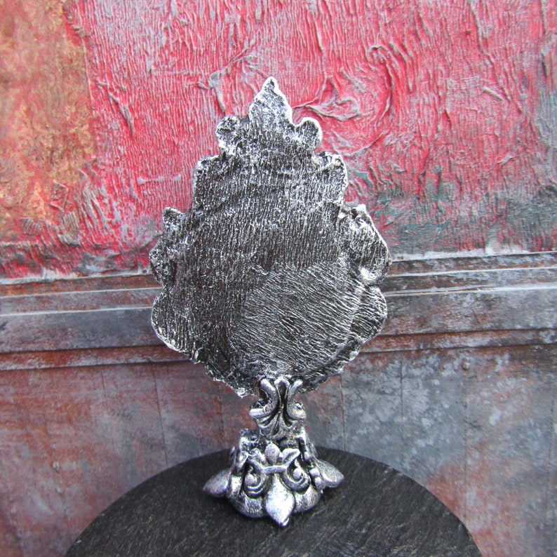 May include: A silver-colored decorative mirror with an ornate, tree-like design. The mirror is standing on a black surface.