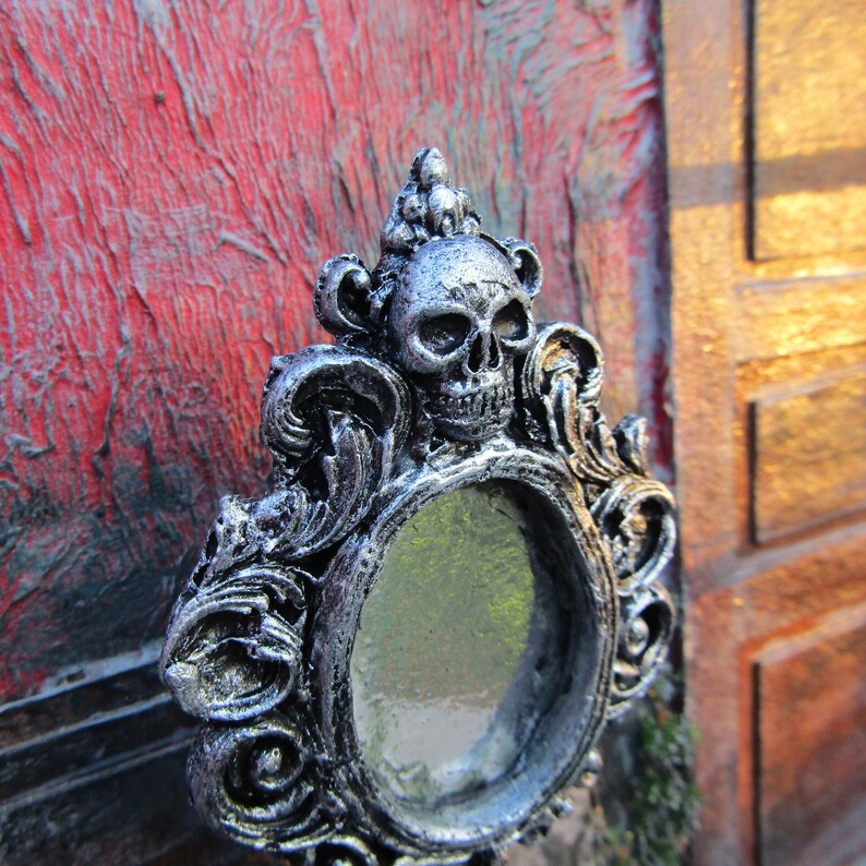 May include: A silver ornate mirror with a skull design. The mirror is round and has a silver frame with intricate details. The skull is silver and has a black eye socket.