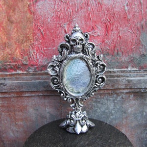 May include: A silver ornate tabletop mirror with a skull design at the top and a round mirror in the center. The mirror is on a black surface.