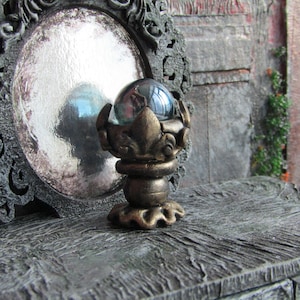 May include: A decorative, ornate, gold-colored stand with a round, clear glass orb on top. The stand has a detailed, intricate design with a floral motif.