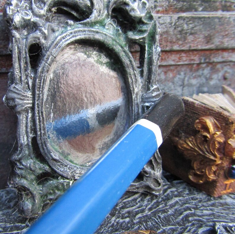 May include: A vintage-style ornate silver-colored metal picture frame with a mirror inside. The mirror is reflecting a blue and white striped pattern. A blue pencil is lying on top of the frame.