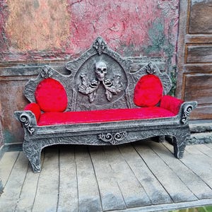 May include: A miniature gothic-style bench with a gray frame and red velvet cushions. The back of the bench features a skull and ornate carvings. The bench is designed with a spooky, Halloween theme.