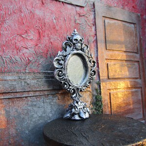 May include: A silver ornate tabletop mirror with a skull design on the top and a blank oval mirror. The mirror is sitting on a dark wooden surface.