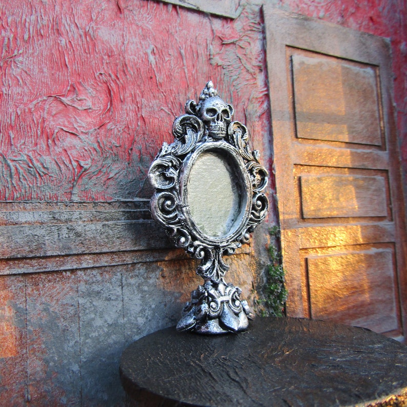 May include: A silver ornate tabletop mirror with a skull design on the top and a blank oval mirror. The mirror is sitting on a dark wooden surface.