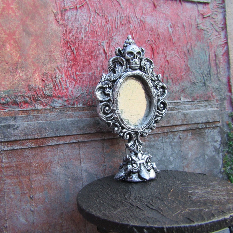 May include: A silver ornate tabletop mirror with a skull design at the top and a blank oval mirror. The mirror is sitting on a dark wooden table.