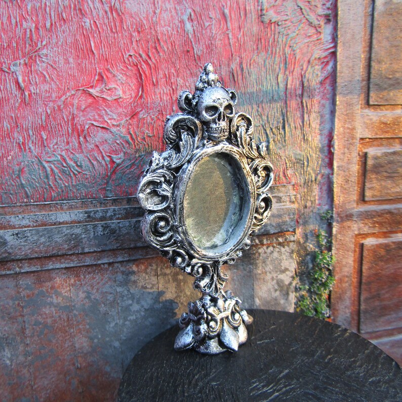 May include: A silver ornate mirror with a skull design and a round mirror. The mirror is on a black surface.