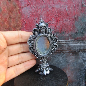 May include: A silver ornate tabletop mirror with a skull design at the top and a round reflective surface. The mirror is on a black surface.