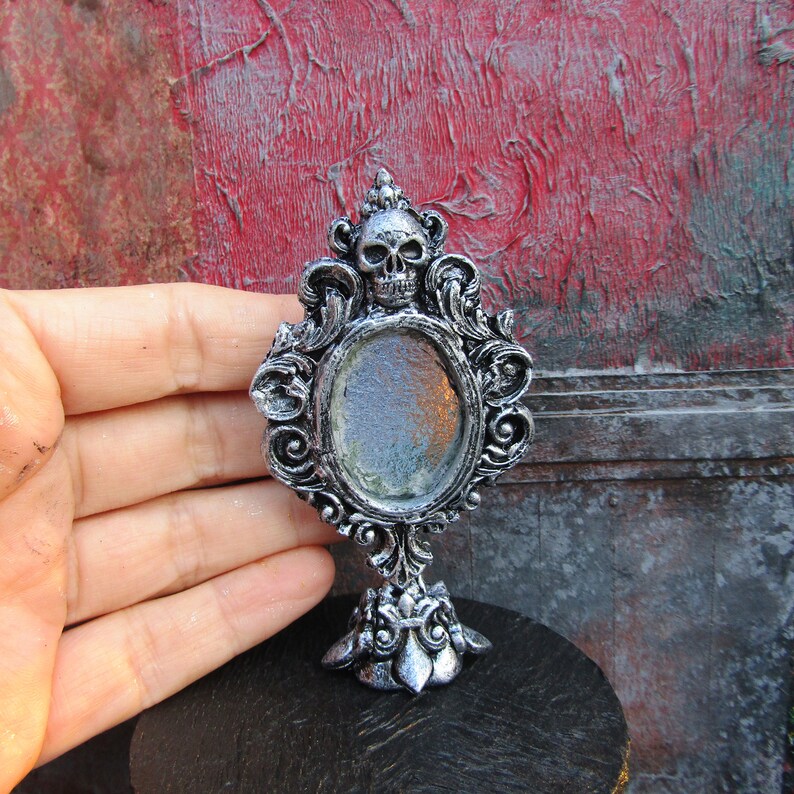 May include: A silver ornate tabletop mirror with a skull design at the top and a round reflective surface. The mirror is on a black surface.