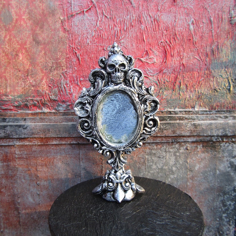 May include: A silver ornate mirror with a skull design at the top. The mirror is standing on a black surface.