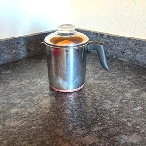 May include: A stainless steel percolator with a cylindrical body, a dark gray handle, and a clear glass knob on the lid. The base of the metal coffee pot features a distinct copper-colored ring, and the surface shows a reflective metallic finish.