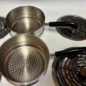 May include: Two stainless steel steamer pots with black handles, each with a perforated bottom. The pots are on a white stove top, next to two electric stove burners.
