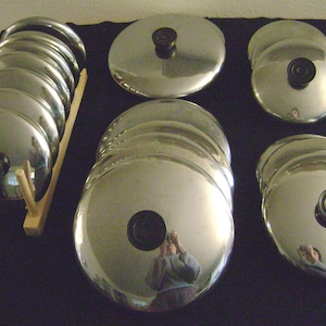 May include: A set of silver-colored metal pot lids with black handles. The lids are stacked and arranged on a wooden stand and on a black surface.