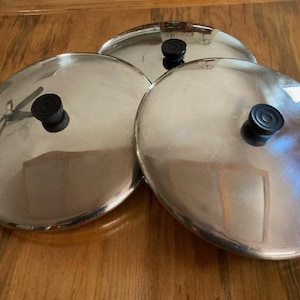 May include: Three stainless steel pot lids with black knobs are arranged on a wooden surface. The lids are circular and reflect the surrounding environment. The knobs are round and made of a dark material.