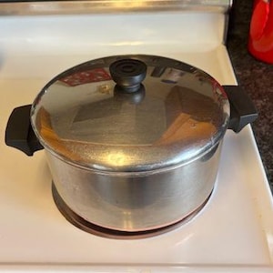 May include: A stainless steel pot with a lid and black handles sits on a white stovetop. The pot has a shiny, reflective surface and a black knob on the lid. The pot is round and appears to be used for cooking.