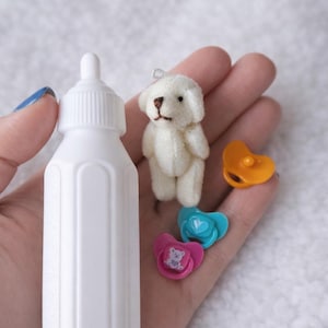 May include: A selection of miniature baby accessories, featuring a white bottle, a small white teddy bear, and three dummies in pink, blue, and orange. The dummies have heart and bear designs. The teddy bear has a brown nose and eyes.