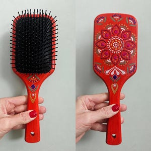 May include: Red wooden hairbrush with a black bristle pad. The handle is decorated with a floral mandala design in shades of pink, white, and blue. The brush is held by a person.