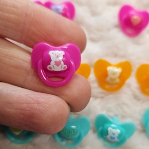 May include: A pink dummy with a white bear design and a heart on the bear's chest. The dummy is made of plastic and has a heart-shaped ring.
