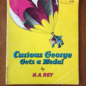 May include: A vintage children's book titled "Curious George Gets a Medal" by H.A. Rey. The cover is yellow with a graphic of George in a spacesuit descending from a pink and gray parachute. The book has a price tag.