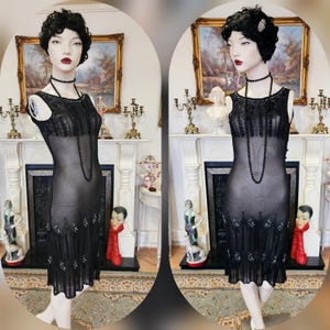 Può includere: Un abito da flapper nero con perline e un'overlay trasparente. L'abito ha una scollatura rotonda e una silhouette aderente. L'abito è adornato con perline e paillettes nere.